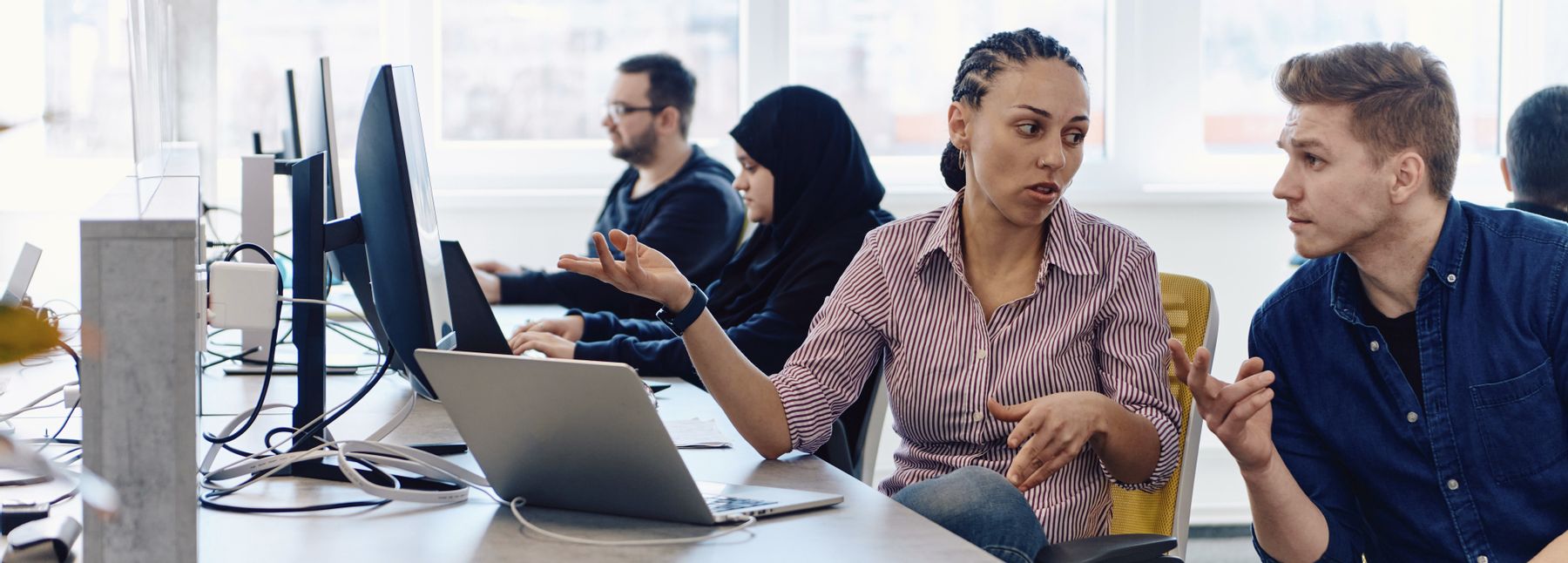 Ein Arbeitsplatz mit Laptop und Stand-PC; im Vordergrund sprechen eine Frau und ein Mann sitzend am Schreibtisch, im Hintergrund sitzen weitere Frau und Mann.