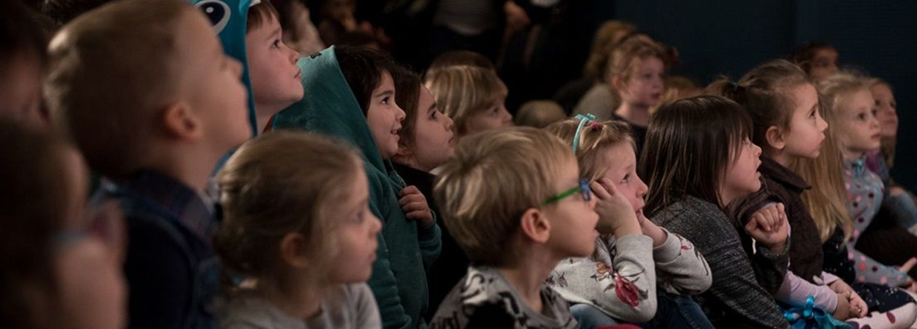 Mehrere Kinder schauen gespannt auf eine Bühne, die sich außerhalb des Bildes befindet