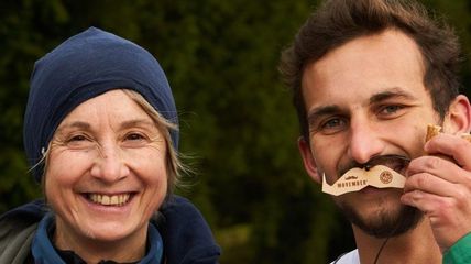 Zwei Personen, eine hält einen Holzschnurrbart vors Gesicht